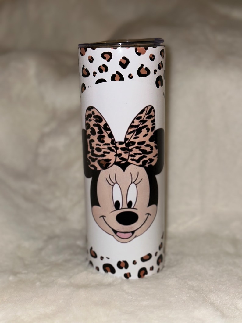 May include: A white tumbler featuring Minnie Mouse with a leopard print bow and accents. The tumbler has a stainless steel appearance with a lid. The design includes a leopard print pattern around the top and bottom.