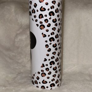 May include: A stainless steel tumbler with a black lid. The tumbler features a white background with a leopard print pattern in brown and black spots. A black circle is visible on the white background.