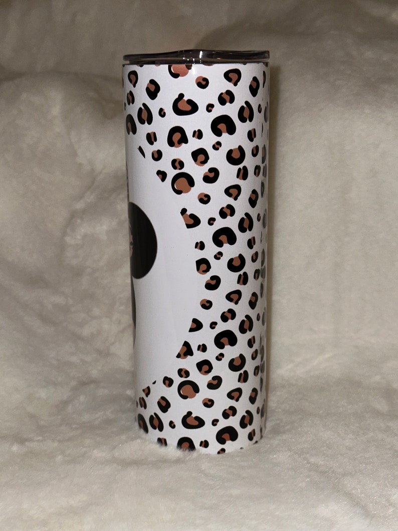 May include: A stainless steel tumbler with a black lid. The tumbler features a white background with a leopard print pattern in brown and black spots. A black circle is visible on the white background.
