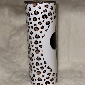 May include: A stainless steel tumbler with a white background and a leopard print pattern in brown and black. The tumbler has a black circle with white text on it. The lid is silver.