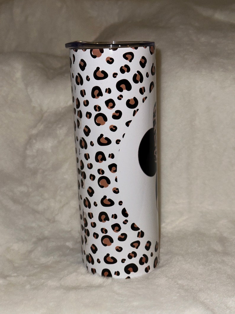 May include: A stainless steel tumbler with a white background and a leopard print pattern in brown and black. The tumbler has a black circle with white text on it. The lid is silver.