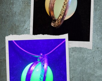 May include: A pendant necklace featuring a glowing orb encased in a decorative silver-toned band. The orb appears pale yellow in normal light and bright green under a black light. The necklace is displayed in two photos.