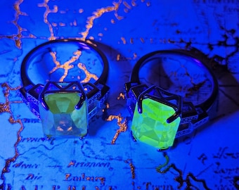 May include: Two rings with large, square, faceted gemstones. One ring has a yellow gem, the other a glowing green gem. The rings are set on a blue map with gold accents. The rings have a dark metal band.