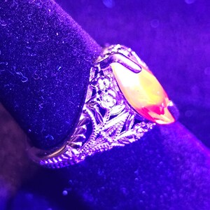 May include: Close-up of a gold-toned ring with an oval, amber-colored stone. The ring features intricate filigree detailing around the stone and band, with small clear stones. The ring is set against a dark, velvety background.