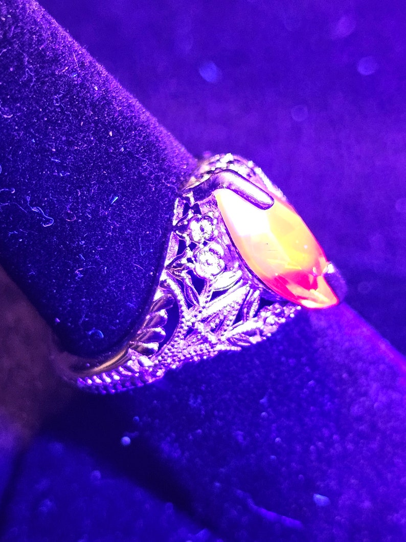 May include: Close-up of a gold-toned ring with an oval, amber-colored stone. The ring features intricate filigree detailing around the stone and band, with small clear stones. The ring is set against a dark, velvety background.