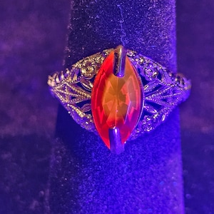 May include: A vintage-style ring with an orange, oval-shaped gemstone. The ring has an ornate, filigree design in a gold tone, with intricate details surrounding the central stone. The ring is displayed on a dark, textured surface.