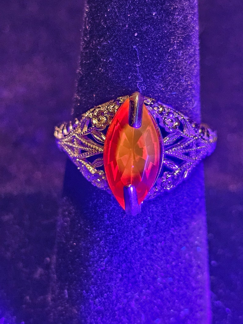 May include: A vintage-style ring with an orange, oval-shaped gemstone. The ring has an ornate, filigree design in a gold tone, with intricate details surrounding the central stone. The ring is displayed on a dark, textured surface.