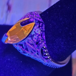 May include: A silver ring with an oval, yellow gemstone. The ring has intricate floral and leaf detailing around the band. The gemstone is set in a prong setting, and the ring is displayed against a dark background.