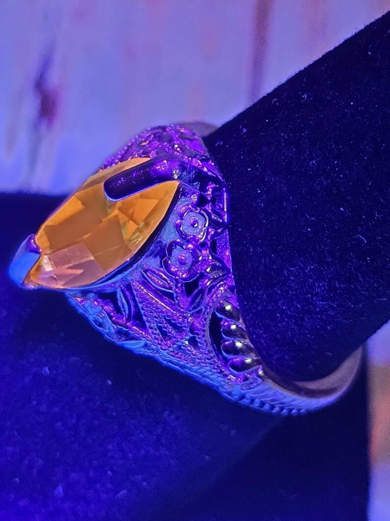 May include: A silver ring with an oval, yellow gemstone. The ring has intricate floral and leaf detailing around the band. The gemstone is set in a prong setting, and the ring is displayed against a dark background.