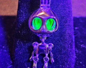 May include: A silver skeleton charm necklace with a skull pendant featuring green eyes. The pendant is hanging from a silver chain.