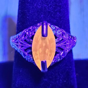 May include: A vintage ring with an oval, yellow gemstone set in a filigree band. The ring's metal has an ornate, floral design. The gemstone is held in place by two dark metal bars. The ring is displayed on a dark blue surface.