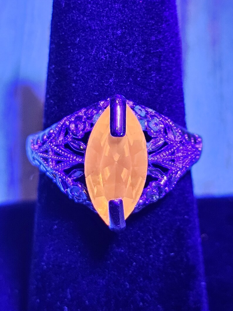 May include: A vintage ring with an oval, yellow gemstone set in a filigree band. The ring's metal has an ornate, floral design. The gemstone is held in place by two dark metal bars. The ring is displayed on a dark blue surface.