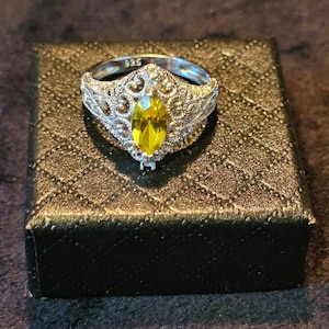 May include: A silver ring with a yellow marquise-shaped gemstone set in a filigree design. The ring is in a black box.