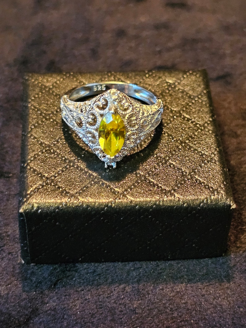 May include: A silver ring with a yellow marquise-shaped gemstone set in a filigree design. The ring is in a black box.