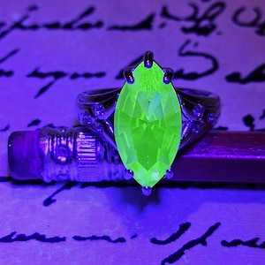 May include: A silver ring with a large, marquise-cut, neon green gemstone. The ring is set against a purple background with script and a pencil.