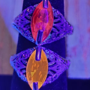 May include: Two ornate rings with filigree detailing. Each ring features a large, marquise-cut gemstone, one orange and one yellow. The rings are set against a dark background, with a purple hue from the lighting.