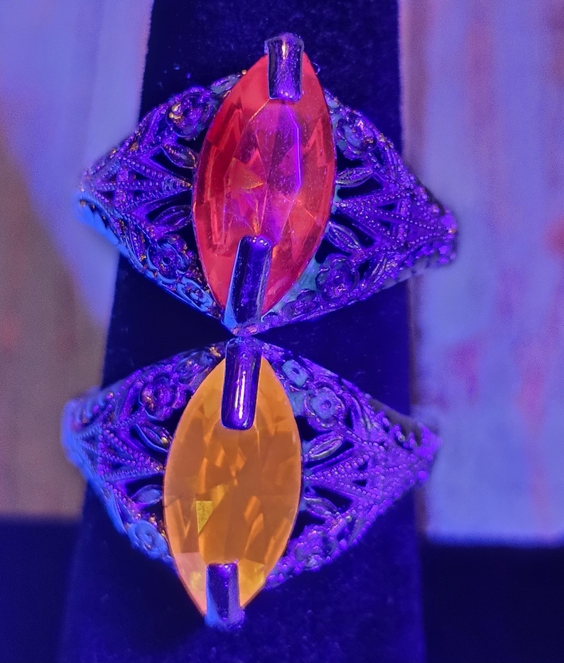 May include: Two ornate rings with filigree detailing. Each ring features a large, marquise-cut gemstone, one orange and one yellow. The rings are set against a dark background, with a purple hue from the lighting.