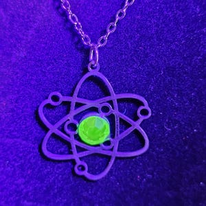 May include: A silver necklace with an atom pendant featuring a green crystal in the centre. The necklace is on a purple background.
