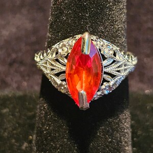 May include: A silver ring with a large, marquise-cut, red gemstone. The ring features intricate filigree detailing around the gemstone and along the band, with small, clear accent stones. The ring is displayed on a black velvet surface.