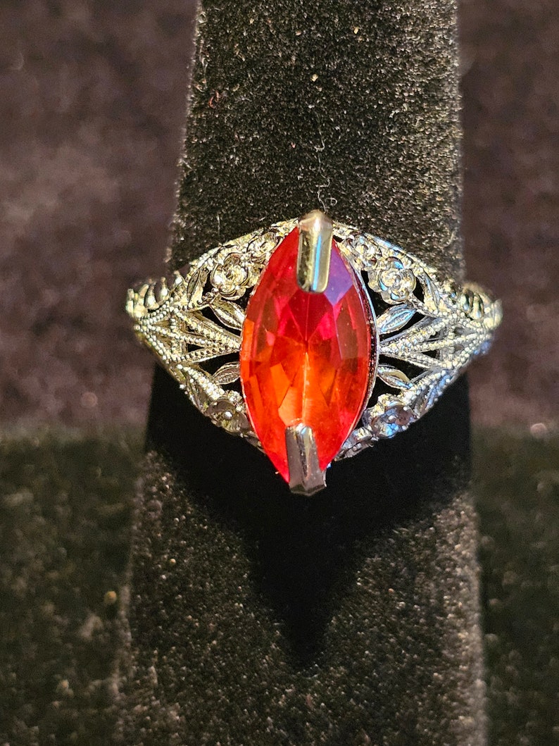 May include: A silver ring with a large, marquise-cut, red gemstone. The ring features intricate filigree detailing around the gemstone and along the band, with small, clear accent stones. The ring is displayed on a black velvet surface.
