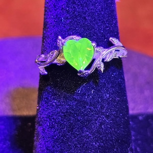May include: A silver-toned ring featuring a heart-shaped, neon green gemstone. The ring has a floral design on the band, with leaves and vines wrapping around the finger. The gemstone is the focal point, catching the light.
