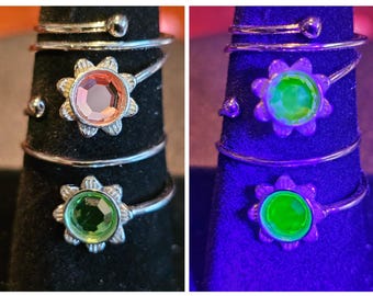 May include: Two silver-toned rings with floral-shaped settings. One ring features a pink gemstone, the other a green gemstone. The rings are displayed on a black surface. The second image shows the rings under a black light.