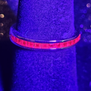 Puede incluir: Un anillo de metal con un engaste de canal continuo de piedras de corte cuadrado que emiten una fluorescencia roja brillante y vibrante bajo luz ultravioleta. La banda tiene un acabado liso y pulido con un ancho uniforme.