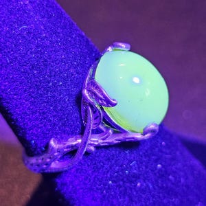 May include: A silver ring with a large, round, green gemstone. The ring is set on a dark blue velvet surface. The gemstone glows brightly under a UV light.