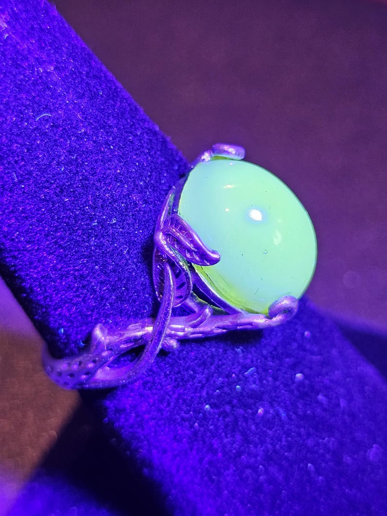May include: A silver ring with a large, round, green gemstone. The ring is set on a dark blue velvet surface. The gemstone glows brightly under a UV light.