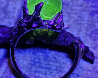 May include: A silver ring featuring a glowing green orb held by a dark, claw-like setting. The ring's band is smooth and polished, contrasting with the textured setting. The orb appears to be translucent, and the overall effect is mystical.