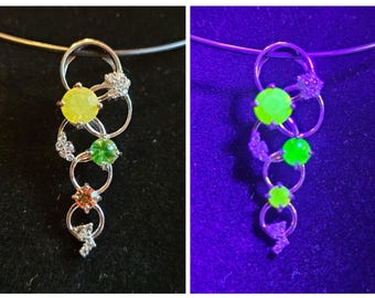 May include: Silver pendant necklace with a series of interlocking circles. The pendant features yellow, green, and orange gemstones, along with small clusters of clear stones. The necklace is shown under normal and UV light.