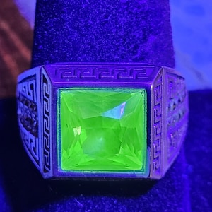 May include: A silver-toned ring featuring a square-cut, vibrant neon green gemstone. The metal band is decorated with a repeating geometric Greek key pattern along the sides and the top border, glowing under ultraviolet light conditions.