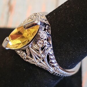 May include: A silver ring with a large, yellow marquise-cut gemstone. The ring features intricate floral and leaf detailing on the band and around the gemstone, with small, clear accent stones. The ring is displayed on a black surface.