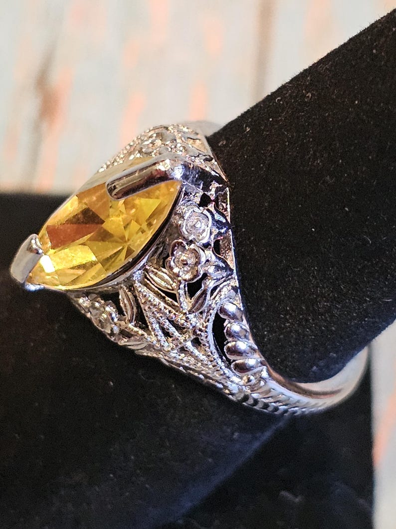 May include: A silver ring with a large, yellow marquise-cut gemstone. The ring features intricate floral and leaf detailing on the band and around the gemstone, with small, clear accent stones. The ring is displayed on a black surface.