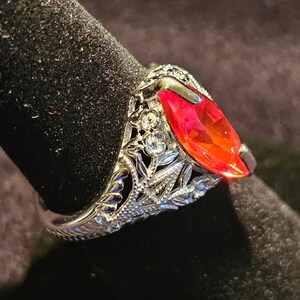 May include: A silver ring with a marquise-cut, vibrant red gemstone. The ring features intricate filigree detailing around the stone and along the band, with small, clear accent stones. The ring is displayed on a black surface.