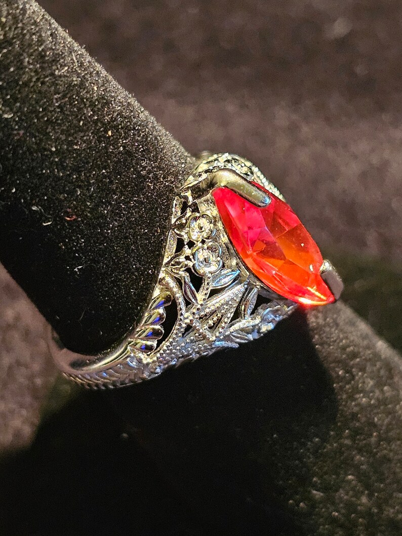 May include: A silver ring with a marquise-cut, vibrant red gemstone. The ring features intricate filigree detailing around the stone and along the band, with small, clear accent stones. The ring is displayed on a black surface.