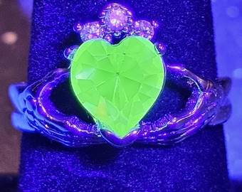 May include: A Claddagh ring with a neon green heart-shaped gemstone. The ring features a crown and hands design, with smaller gemstones around the crown. The ring is set against a dark blue background.