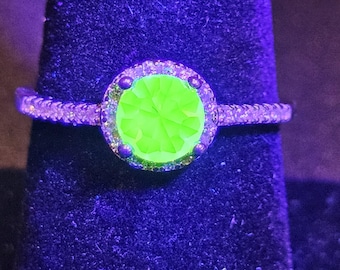 May include: A ring featuring a round, faceted center stone that glows a vibrant neon green under ultraviolet light. The stone is surrounded by a halo of small, clear stones, and the band is encrusted with additional small, clear, sparkling stones.