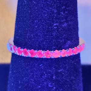 May include: A thin metal ring featuring a continuous row of small, round, vibrant neon pink stones that glow intensely. The band is set with these uniform, circular gems that span the entire circumference of the ring, creating a bright, saturated color.