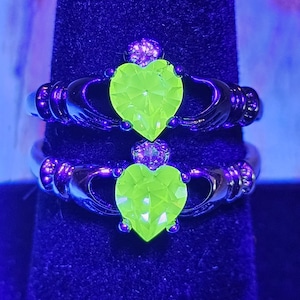 Può includere: Due anelli di Claddagh con fasce nere e pietre centrali a forma di cuore verde neon. Gli anelli presentano mani che stringono il cuore, con una corona sopra. Gli anelli sono esposti su una superficie di velluto nero.