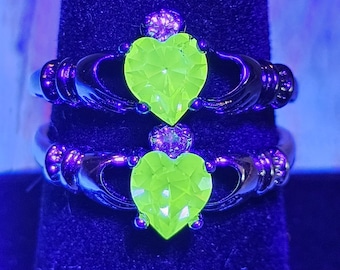 May include: Two Claddagh rings with black bands and neon green heart-shaped center stones. The rings feature hands clasping the heart, with a crown above. The rings are displayed on a black velvet surface.