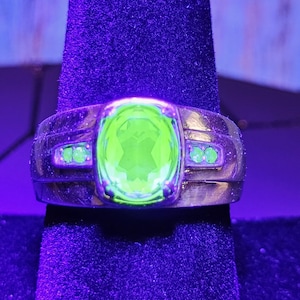 May include: A gold ring with a large, oval-shaped green gemstone surrounded by smaller green gemstones. The ring is on a purple velvet surface.