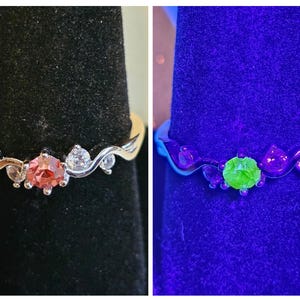 Könnte beinhalten: Ein silberner Ring mit Wellendesign, mit einem zentralen rosa Edelstein und kleineren klaren Steinen. Der Ring wird auf einem schwarzen Samthintergrund präsentiert. Das zweite Bild zeigt den Ring unter Schwarzlicht, wobei der Mittelstein grün leuchtet.