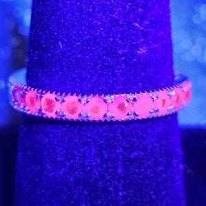 May include: A silver-toned band ring featuring a continuous row of round, faceted stones that glow with a vibrant, neon pink fluorescence under ultraviolet light. The metal setting displays a delicate, textured milgrain edge along the band.