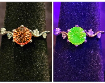 May include: A silver ring with a large orange gemstone in the center and smaller clear gemstones on the band. The ring glows green under a black light.