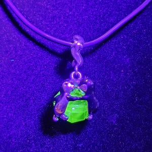 May include: A silver-toned pendant necklace featuring two figures embracing a bright green, faceted gemstone. The pendant hangs from a dark purple cord, set against a dark purple background.