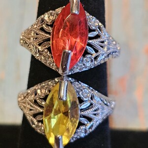 May include: Two rings with marquise-cut gemstones, one red and one yellow, are displayed. The rings have ornate, silver-colored filigree bands with floral and leaf patterns. The gemstones are set with silver-colored prongs.
