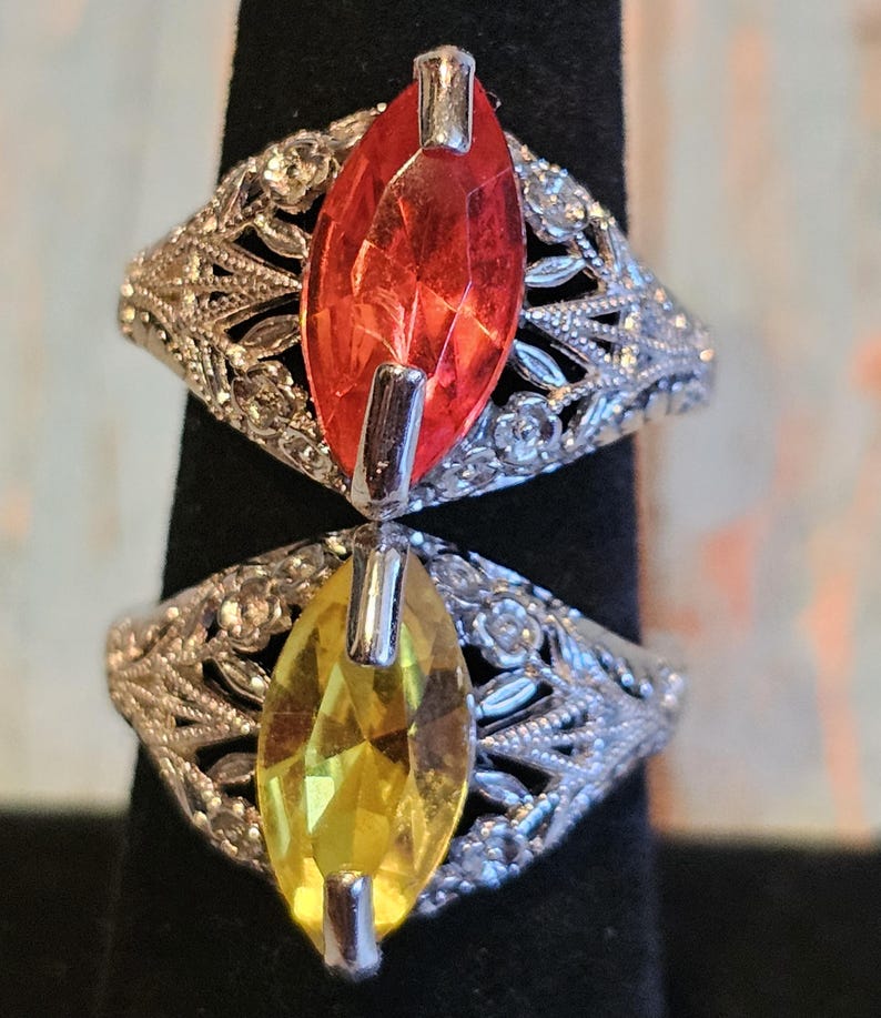 May include: Two rings with marquise-cut gemstones, one red and one yellow, are displayed. The rings have ornate, silver-colored filigree bands with floral and leaf patterns. The gemstones are set with silver-colored prongs.