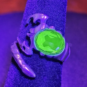 May include: A silver-toned ring with a scorpion design, featuring a large, faceted, neon green gemstone. The ring is displayed on a dark blue velvet surface, illuminated by a purple light.