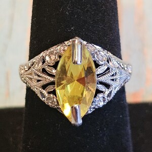 May include: A silver ring with a large, marquise-cut yellow gemstone. The ring features an ornate, filigree design with floral accents around the gemstone. The ring is displayed on a black stand.